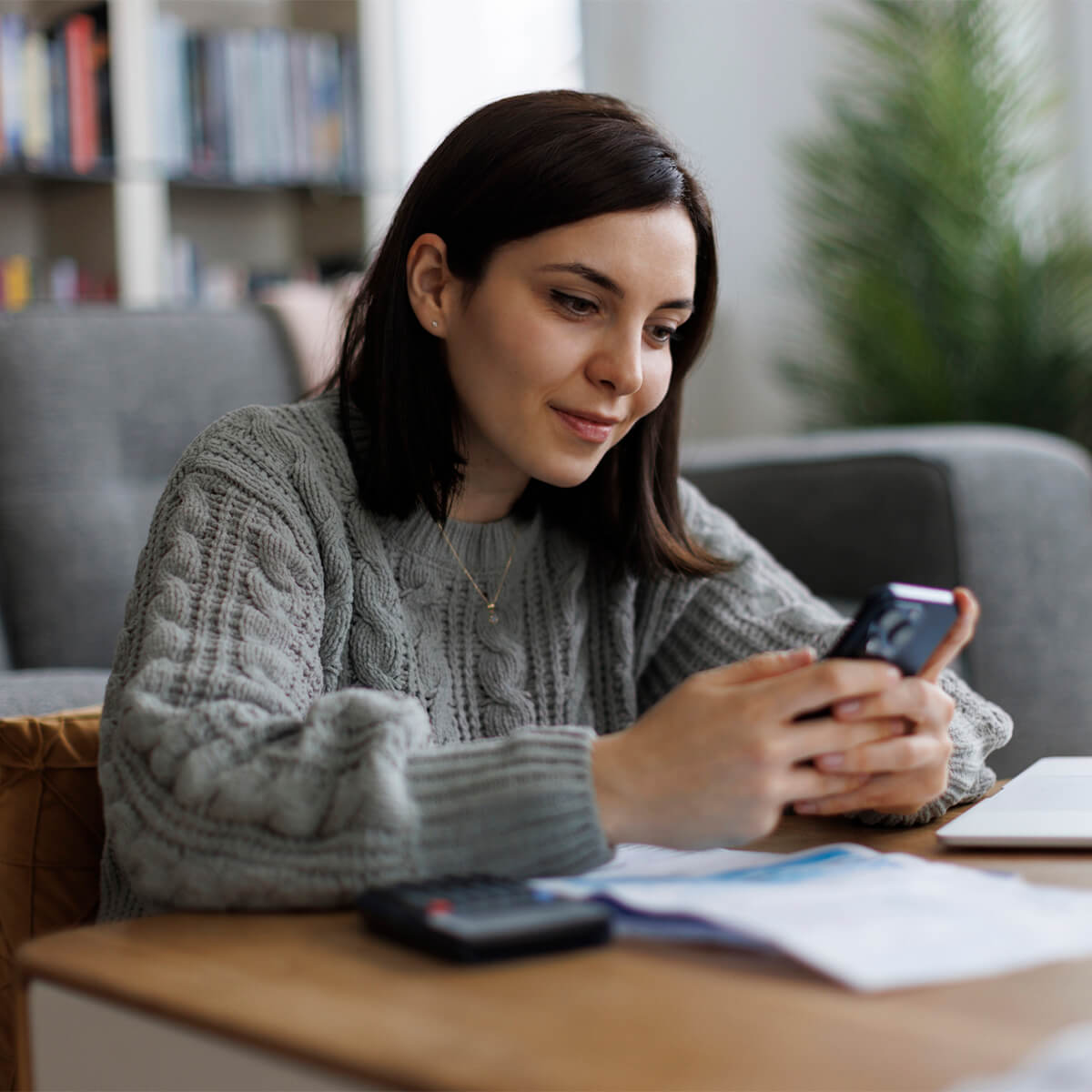 A woman holds a phone while bills lie on the table in front of her.