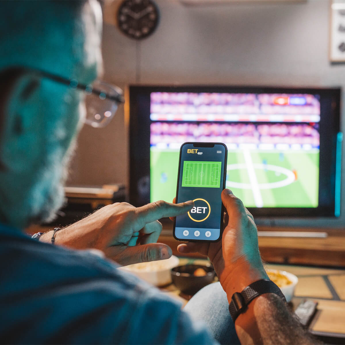 A man watching soccer on TV places a bet using his phone