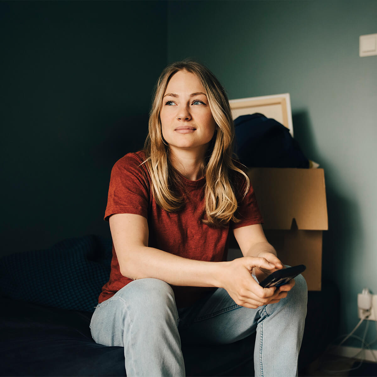 Pensif jeune femme regardant ailleurs tout en étant assis avec un téléphone intelligent dans la chambre