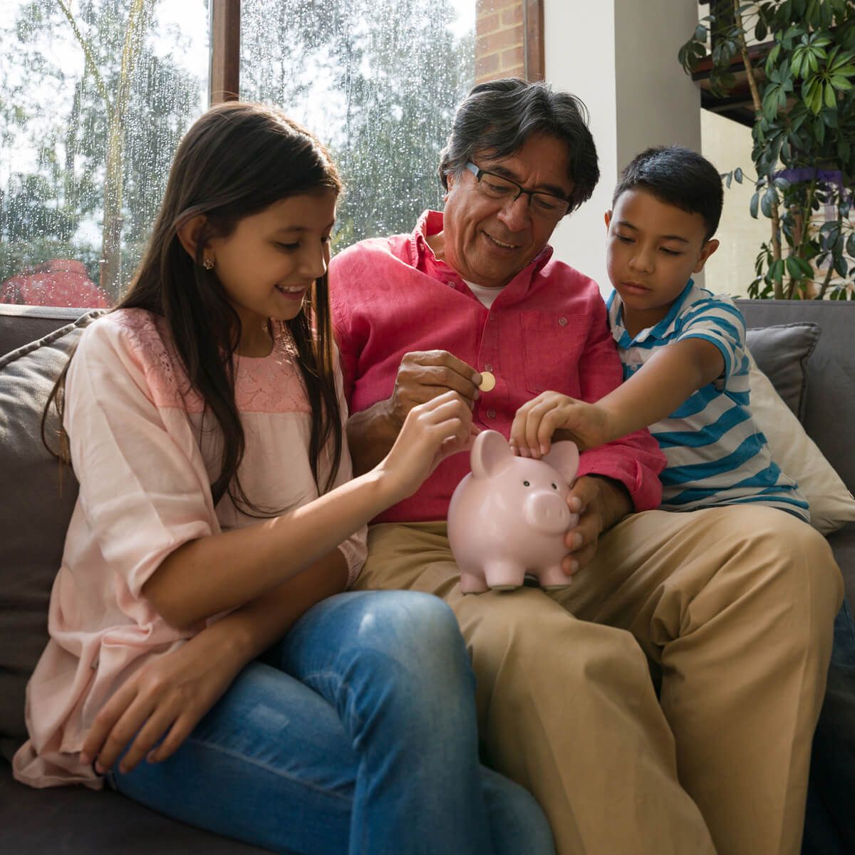 Parent sitting on couch with son and daughter putting money in a piggy bank