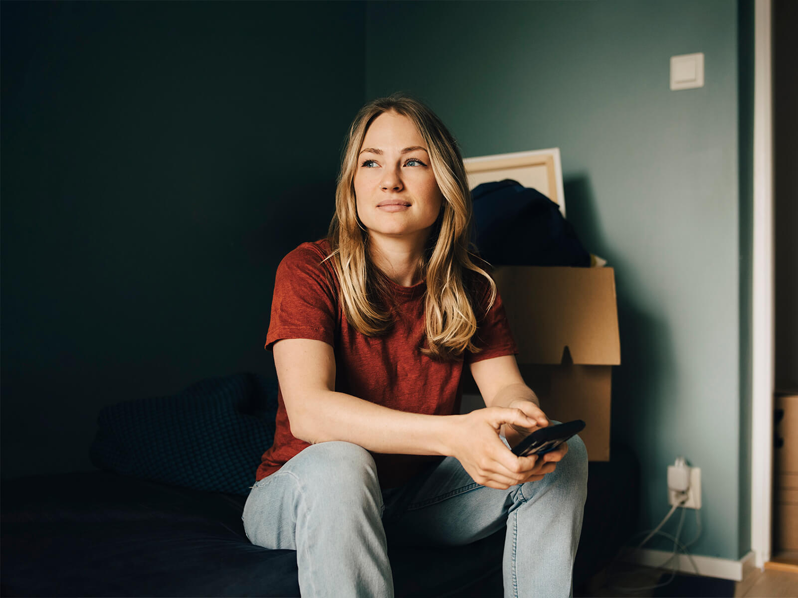 Thoughtful young woman looking away while sitting with smart phone in bedroom - stock photo